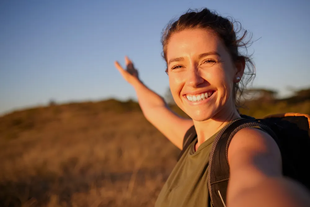 A woman smiles as she takes a selfie with her arm outstretched while on a hike.